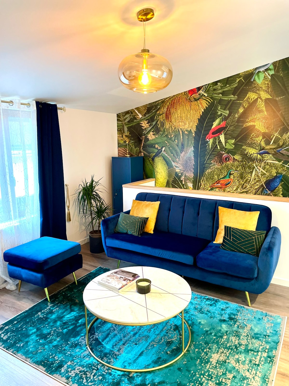 A stylish living area features a blue velvet sofa adorned with decorative cushions and matching ottoman. The space is highlighted by a vibrant botanical wall mural and is completed with a round coffee table atop a textured rug, with natural light filtering through the sheer curtains.