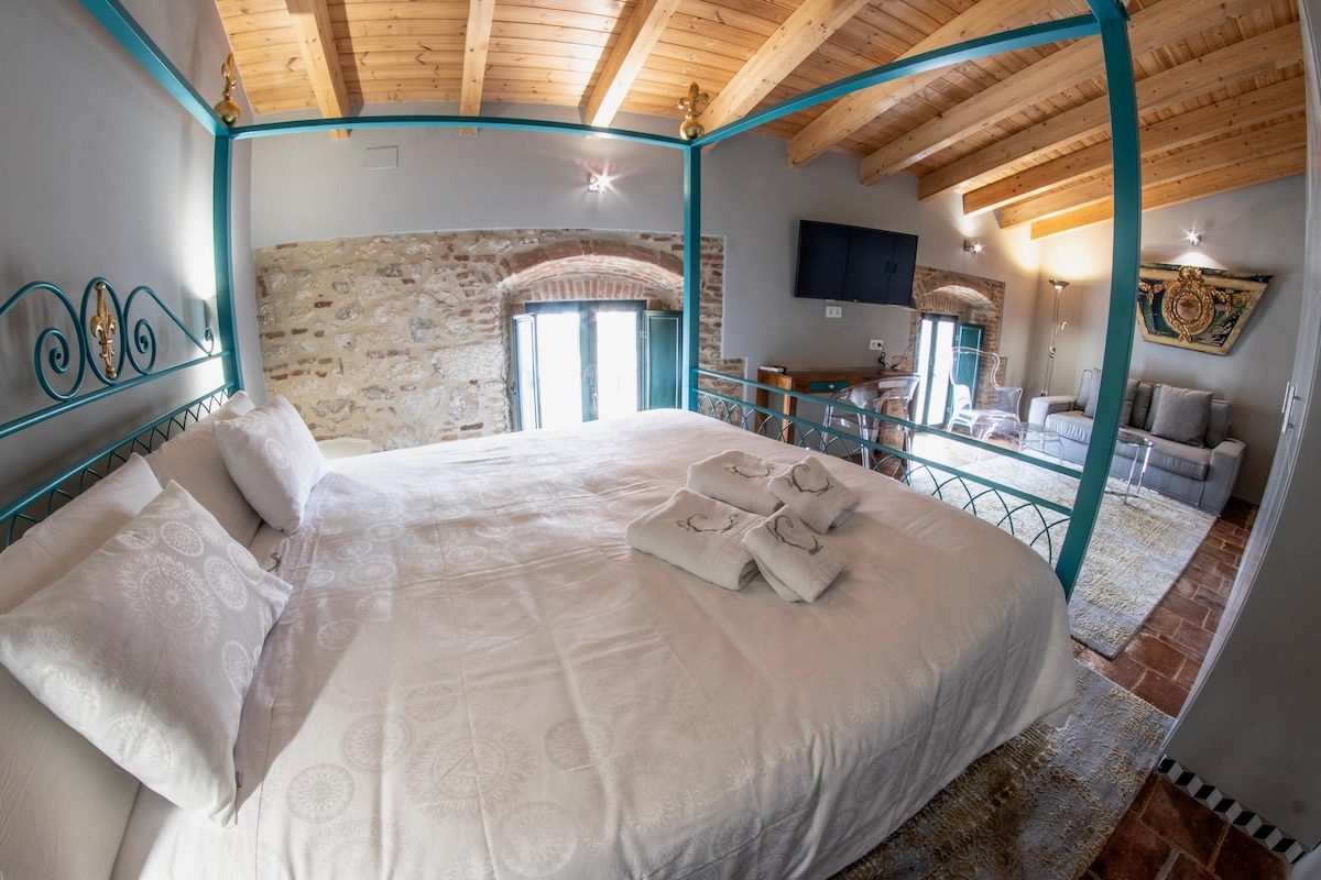 A spacious sleeping area features a large bed with crisp, white linens and neatly arranged towels. Exposed wooden beams enhance the rustic charm, while large windows provide ample natural light. A comfortable seating area is visible in the background, complemented by elegant wall art.