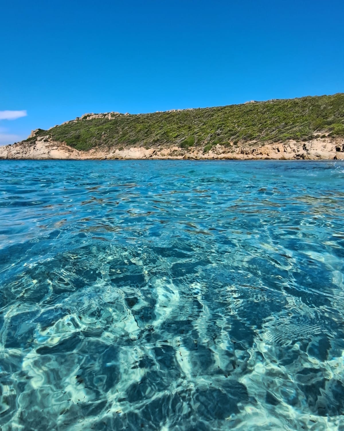 Crystal-clear waters reflect the sunlight, revealing underwater textures and colors. A rugged coastline with lush greenery is visible in the background, contributing to the serene coastal scene under a clear blue sky.
