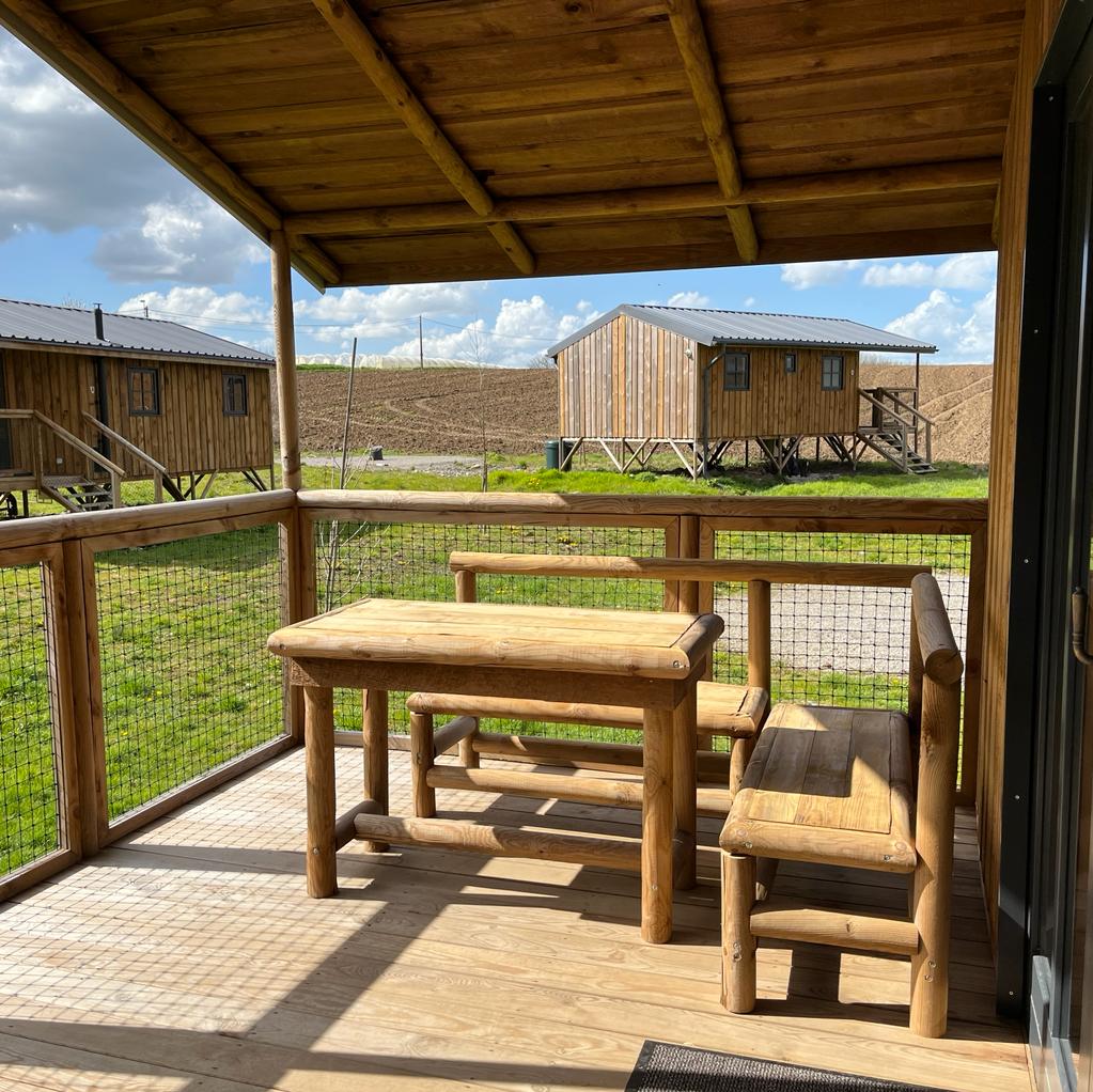 A covered terrace features a wooden table and two benches, designed for outdoor dining. The surrounding area includes grassy landscapes and wooden lodges positioned in the background, providing a natural and tranquil setting.