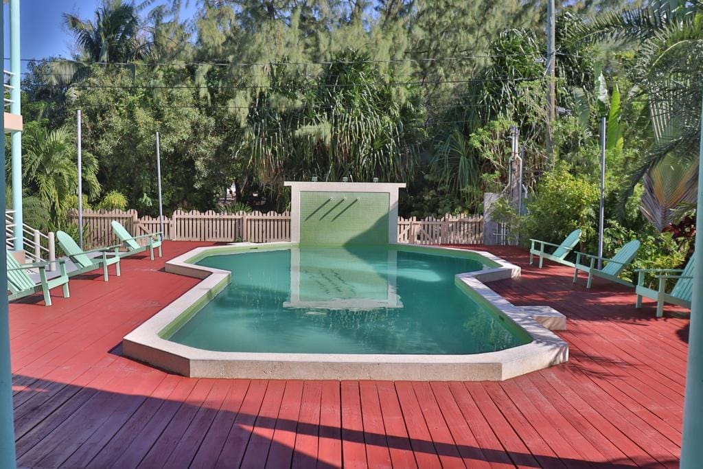 A pool area is depicted with a uniquely shaped pool surrounded by a wooden deck in shades of red. Lounge chairs are arranged around the pool, and lush greenery frames the space, providing a serene setting for relaxation.
