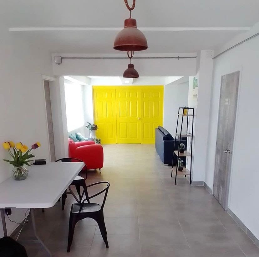 A bright, inviting space is showcased, featuring a vivid yellow wall that adds a pop of color. A comfortable red sofa and a dark blue couch are arranged with a dining table and black chairs. Natural light fills the room from large windows.