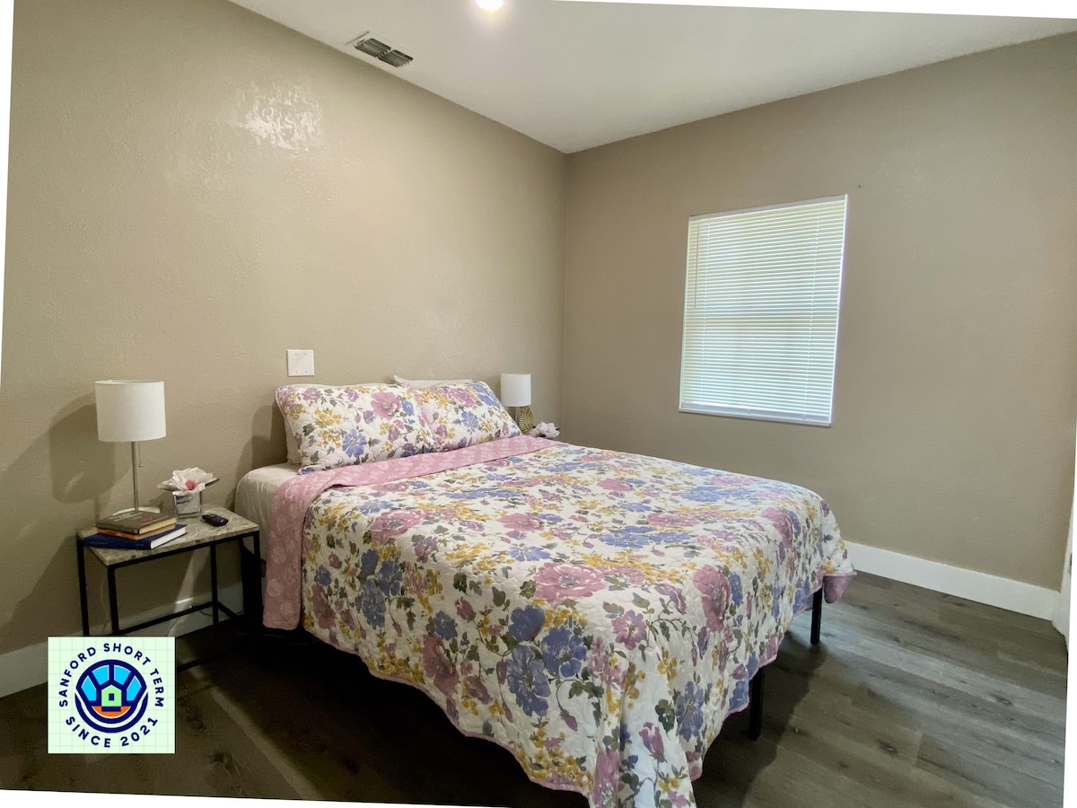 A spacious bedroom is furnished with a queen bed covered in a floral patterned blanket. Two bedside tables hold lamps, and natural light enters through a window with light-colored blinds. The walls are painted a soft neutral tone.