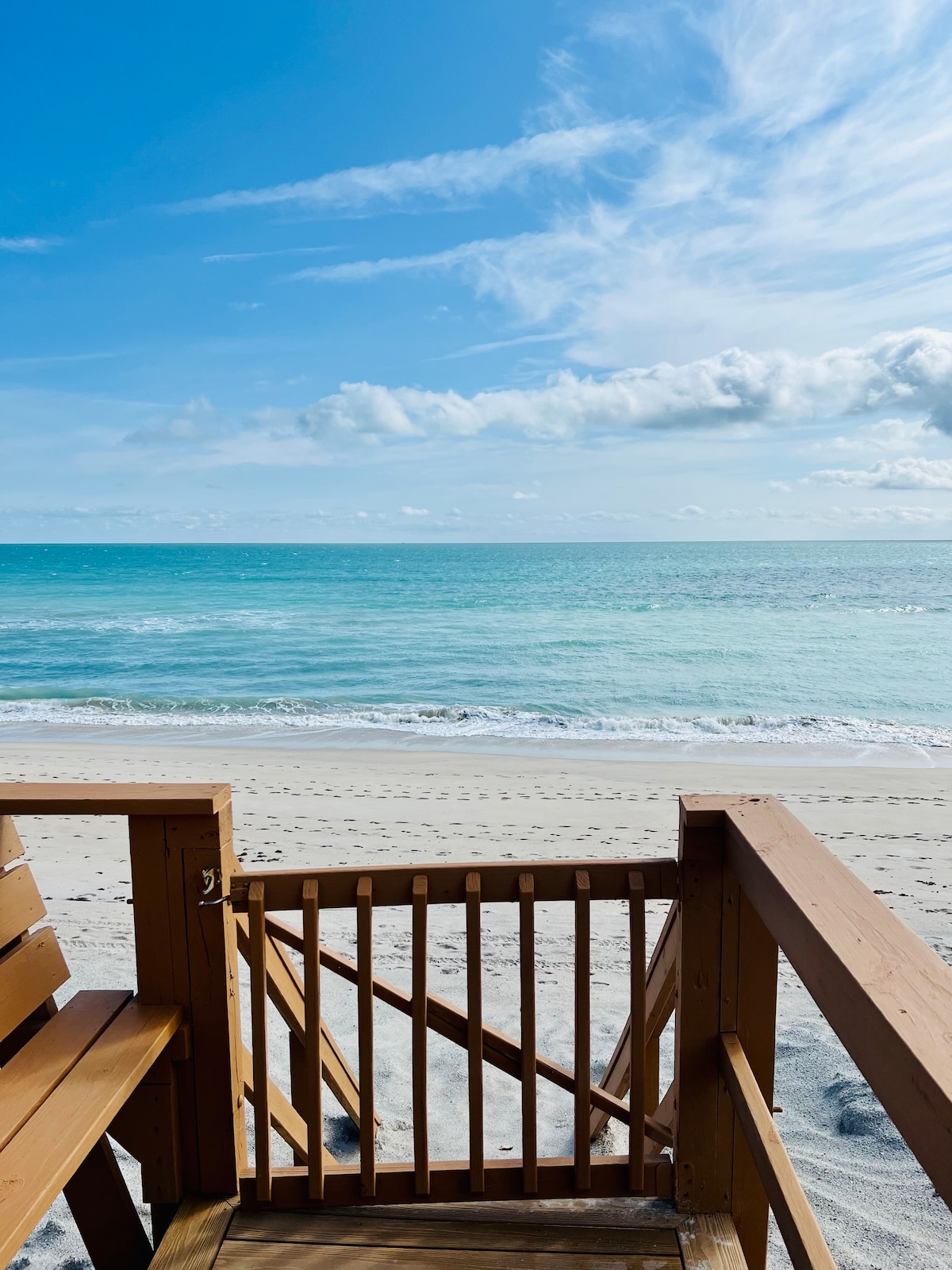 A wooden deck features seating overlooking a serene beach. The vibrant blue ocean stretches towards a clear sky, while gentle waves lap against the shore. Footprints are seen in the sand, indicating a peaceful and inviting coastal atmosphere.