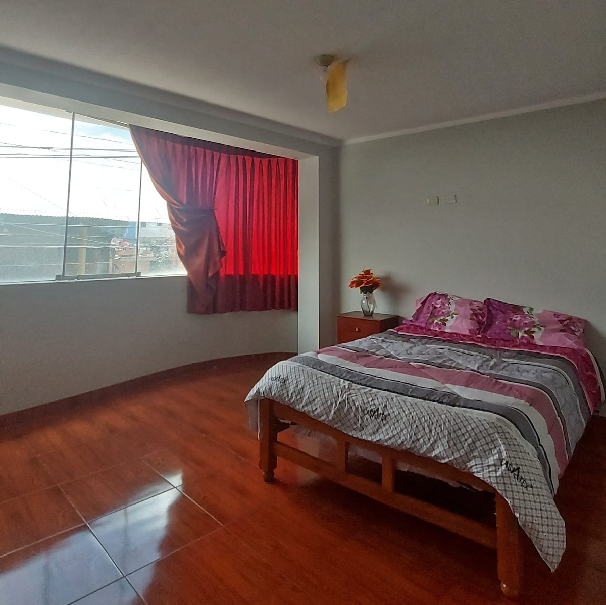 A well-lit bedroom features a spacious bed with a patterned bedspread. A large window adorned with red curtains allows natural light to enter. A bedside table holds a small vase with flowers, complementing the warm wooden floor.