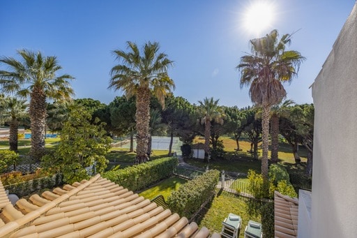 A view from the upper level captures lush palm trees and manicured greenery in the surrounding area. A clear blue sky is visible overhead, with sunlight streaming down, illuminating the outdoor space and hinting at the tranquility of the private garden below.