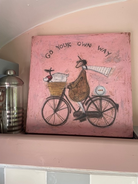 A whimsical painting is elevated on a shelf, featuring a bicycle with a basket carrying a sheep. The background is a soft pink, and text reading 'GO YOUR OWN WAY' curves above the bicycle, adding a playful touch to the interior.