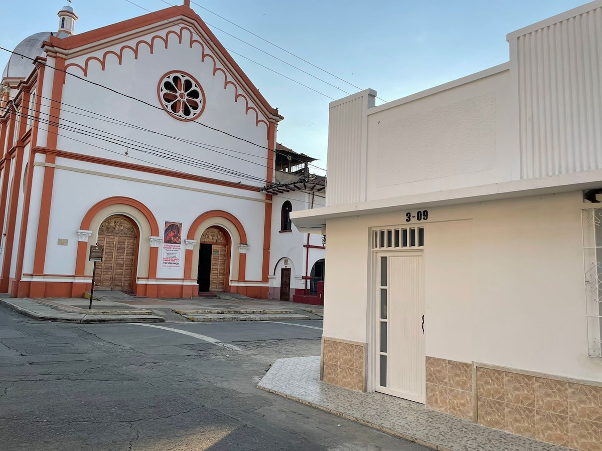 A white building with a minimalist design stands at the corner of a street, featuring a door marked with the number 3-09. A historical church with bright red and orange accents is visible across the street, contrasting with the simplicity of the nearby structure.