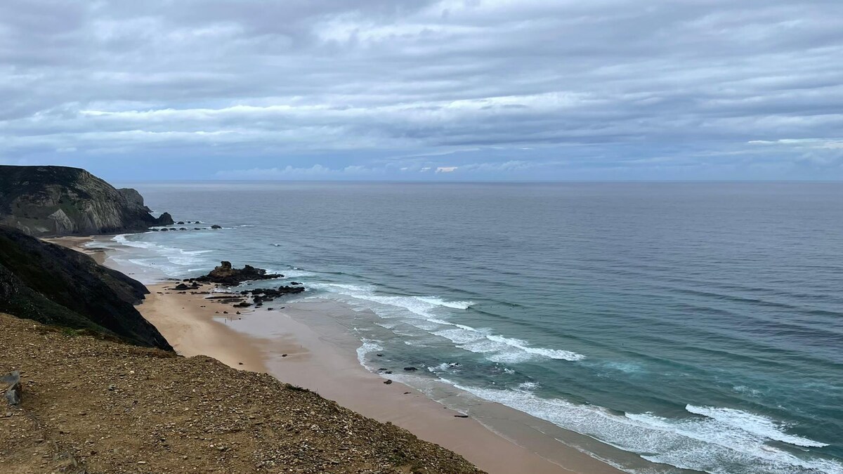 The image showcases a serene beach scene with gentle waves lapping at the shore. Cliffs rise in the background, framing the sandy coastline. The sky is partially cloudy, with shades of grey and hints of blue, reflecting a tranquil coastal atmosphere.