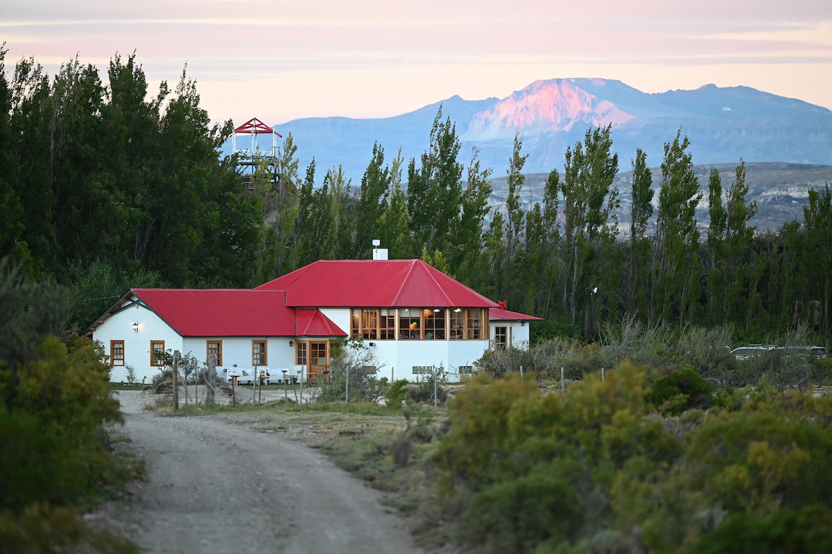 A spacious house with a red roof is nestled among greenery, providing views of distant mountains under soft evening light. A dirt path leads toward the entrance, enhancing the serene landscape of the property.
