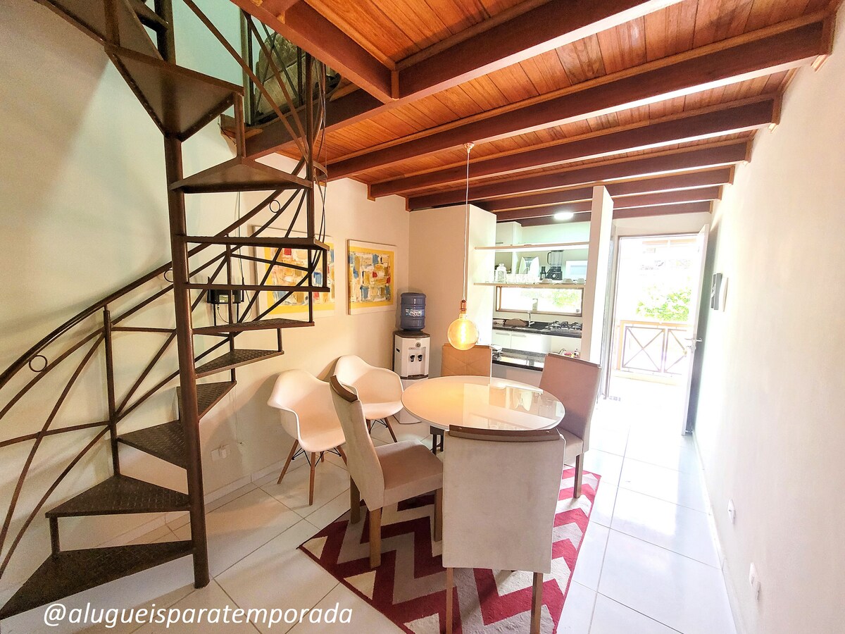 A dining area features a round table surrounded by four light-colored chairs. A spiral staircase leads to an upper level. The kitchen is visible in the background, showcasing modern appliances and cabinetry. Soft natural light enters through sliding glass doors that open to a patio.