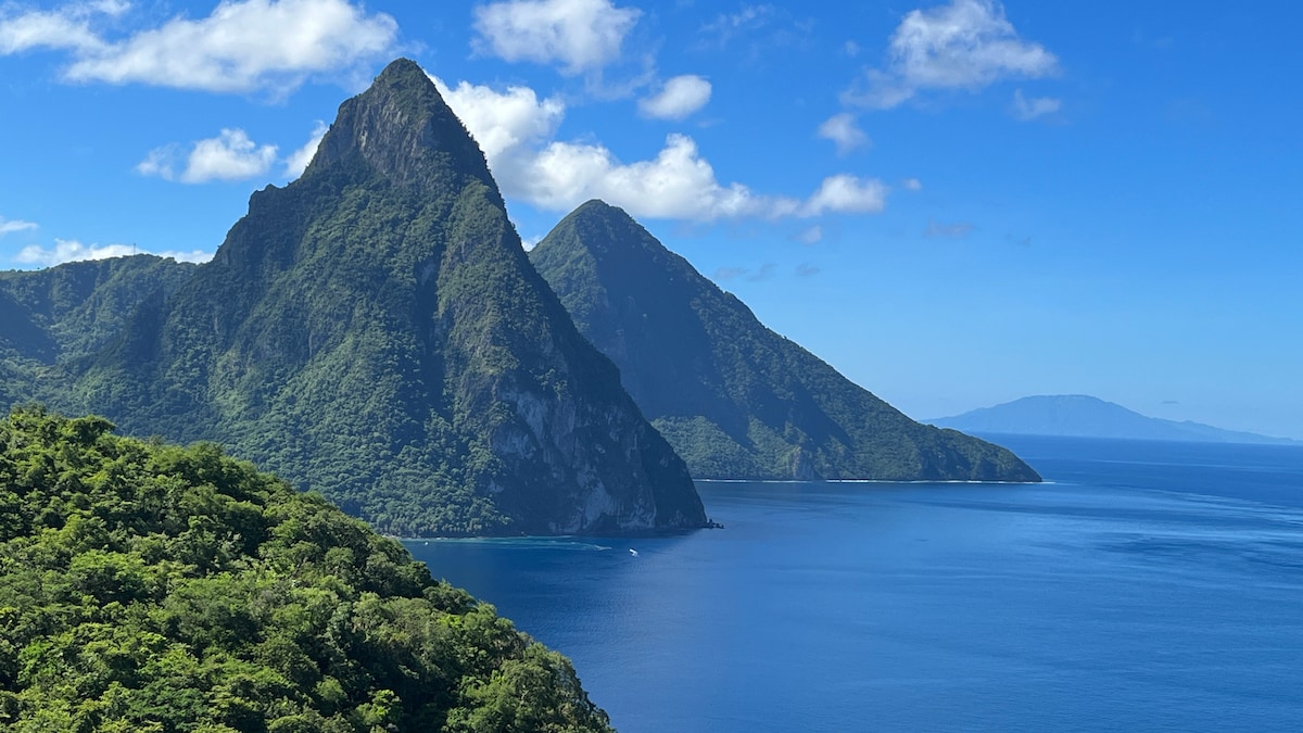 A panoramic view of lush green mountains rises dramatically from the deep blue Caribbean Sea. The iconic Pitons, characterized by their steep, pointed peaks, are framed by a clear sky dotted with a few fluffy clouds.