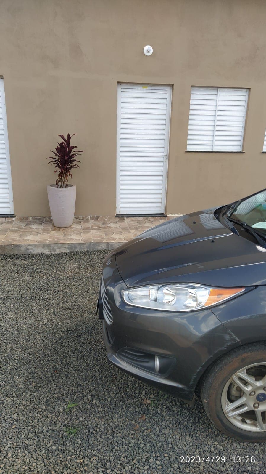 A parked car is positioned in front of a building, with its front partially visible. Two doors can be seen behind the car, featuring white horizontal slats. A potted plant adds a touch of greenery to the entrance area, set against a neutral exterior wall.