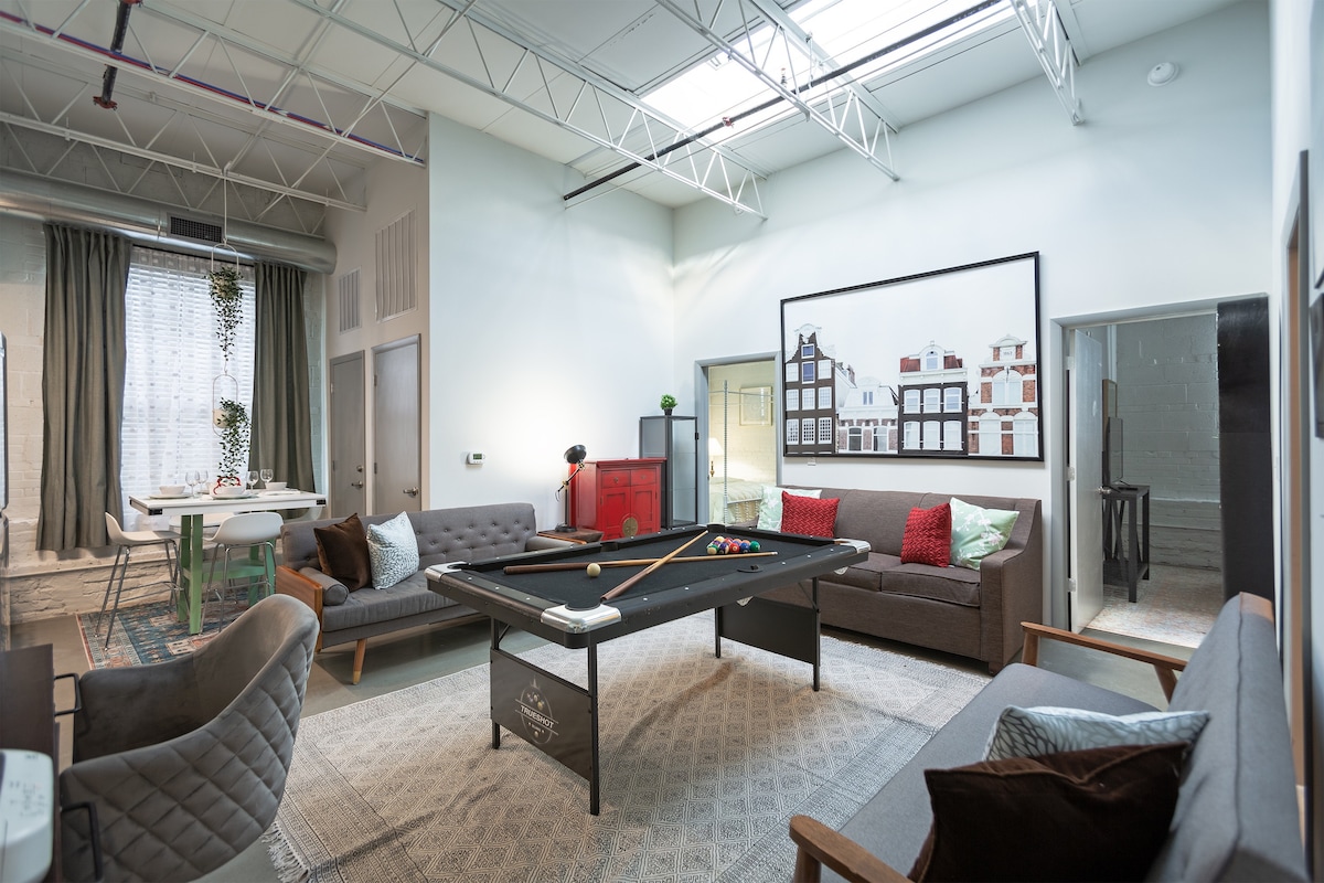 A spacious living area features a foldable pool table at its center, surrounded by two sofas and a chair. Large windows allow natural light to fill the room, while artwork adorns the walls. A dining area is visible in the background, creating a versatile space.