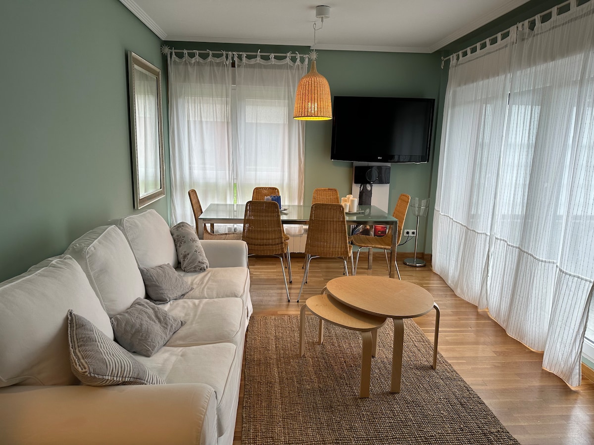 An open living area is presented, featuring a light-colored sofa accompanied by decorative pillows. A round coffee table sits in front, and a dining table is visible with several wicker chairs. Natural light filters through sheer curtains that frame the windows.