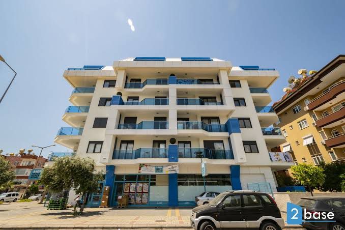 The modern building presents a clean, white exterior with blue accents. Multiple balconies feature glass railings on each floor. The ground level includes commercial spaces, while a row of trees line the sidewalk beside parked vehicles, creating an organized urban setting.