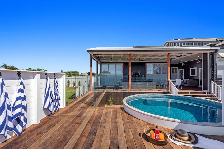 The image shows a modern outdoor deck area featuring a small round pool with a glass fence and wooden flooring. Blue and white striped towels hang on hooks along a white wall. The covered seating area in the background has comfortable outdoor furniture, providing a relaxing space. The open design allows for easy movement between the pool area and the house, giving it a coastal, luxurious vibe perfect for enjoying warm weather. The clear blue sky adds to the bright and inviting atmosphere of the space.