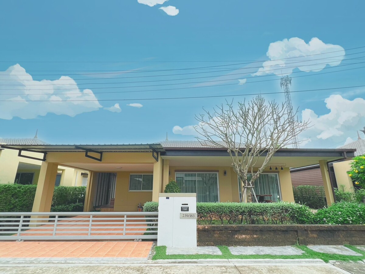 A single-story house with a welcoming porch features large windows framed by greenery. A white mailbox stands prominently at the entrance, while a blossoming tree provides shade. The clear blue sky enhances the overall tranquility of the property, inviting relaxation.