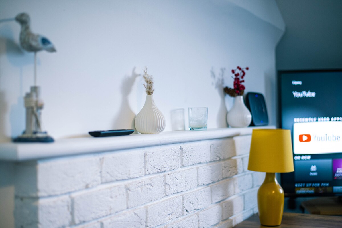 A shelf is adorned with decorative items, including a small bird figurine, a white vase with dried flowers, and a glass. A yellow lamp adds a pop of color, while a smart TV displays the YouTube interface in the background.