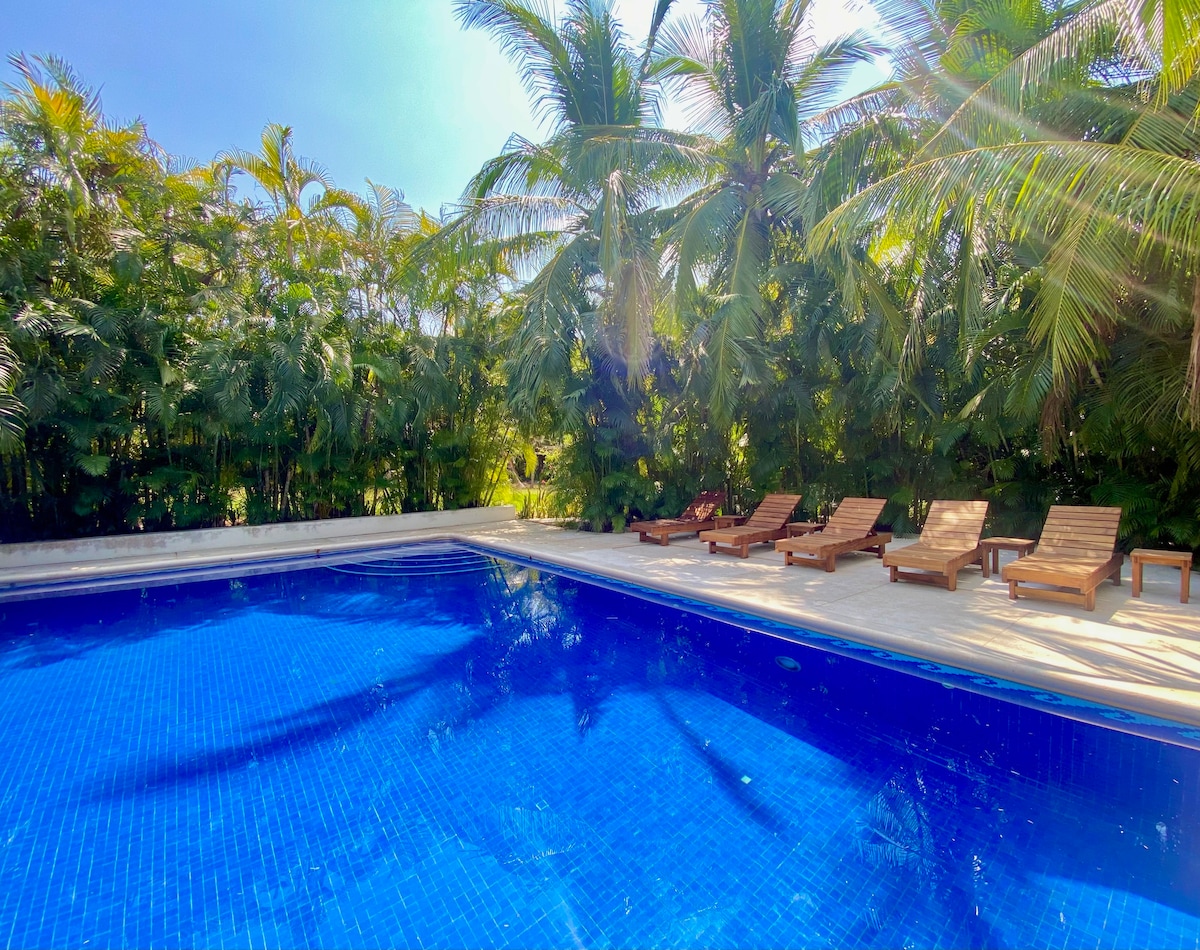A swimming pool surrounded by lush palm trees is featured, with several wooden lounge chairs positioned along the poolside. The water reflects the vibrant blue sky, creating a serene and inviting space for relaxation.