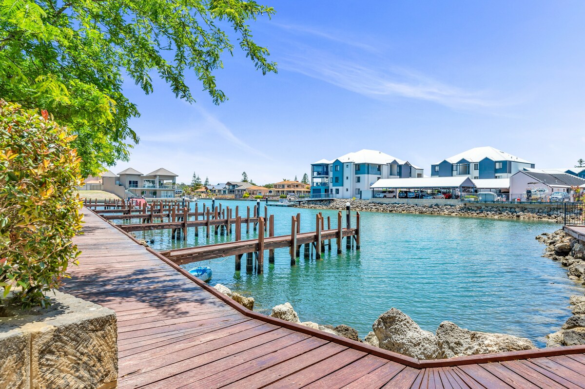 A wooden boardwalk leads to a calm waterway, with moorings visible along the edge. Buildings with light-colored facades are situated across the water, and boats can be seen moored nearby. Lush greenery partially frames the scene, providing a serene backdrop.