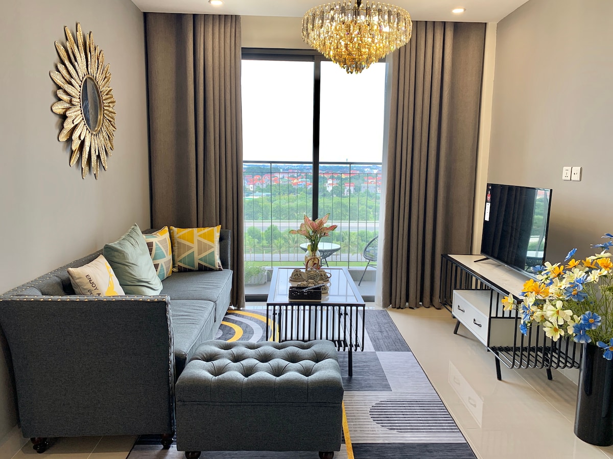 A modern living area features a grey sofa adorned with decorative cushions, complemented by a tufted ottoman. A flat-screen TV is positioned on a low white cabinet. Natural light enters through large glass doors, enhancing the view of the greenery outside. A decorative chandelier hangs above.