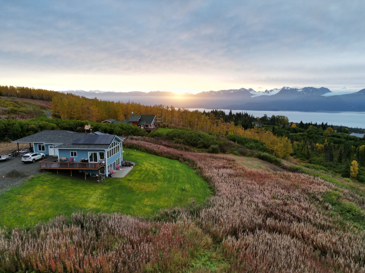 An aerial view captures a modern home set amidst lush green lawns and vibrant fireweed fields, with mountains and water visible in the background. A soft sunrise lights the scene, highlighting the surrounding natural beauty and creating a serene atmosphere.