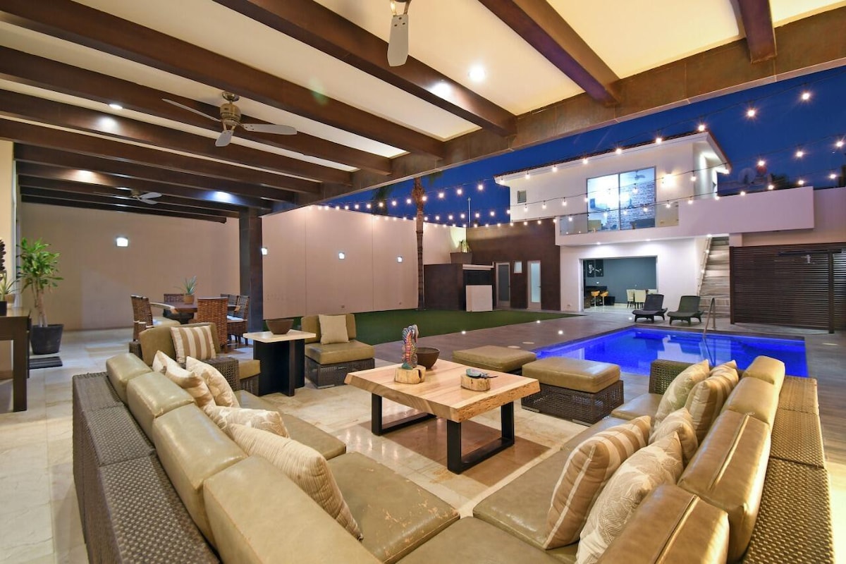 An outdoor lounge area is featured, showcasing a spacious arrangement of comfortable seating around a central table. A swimming pool is observed in the background, while soft lighting and decorative string lights create a serene ambiance. A dining area is visible to one side.