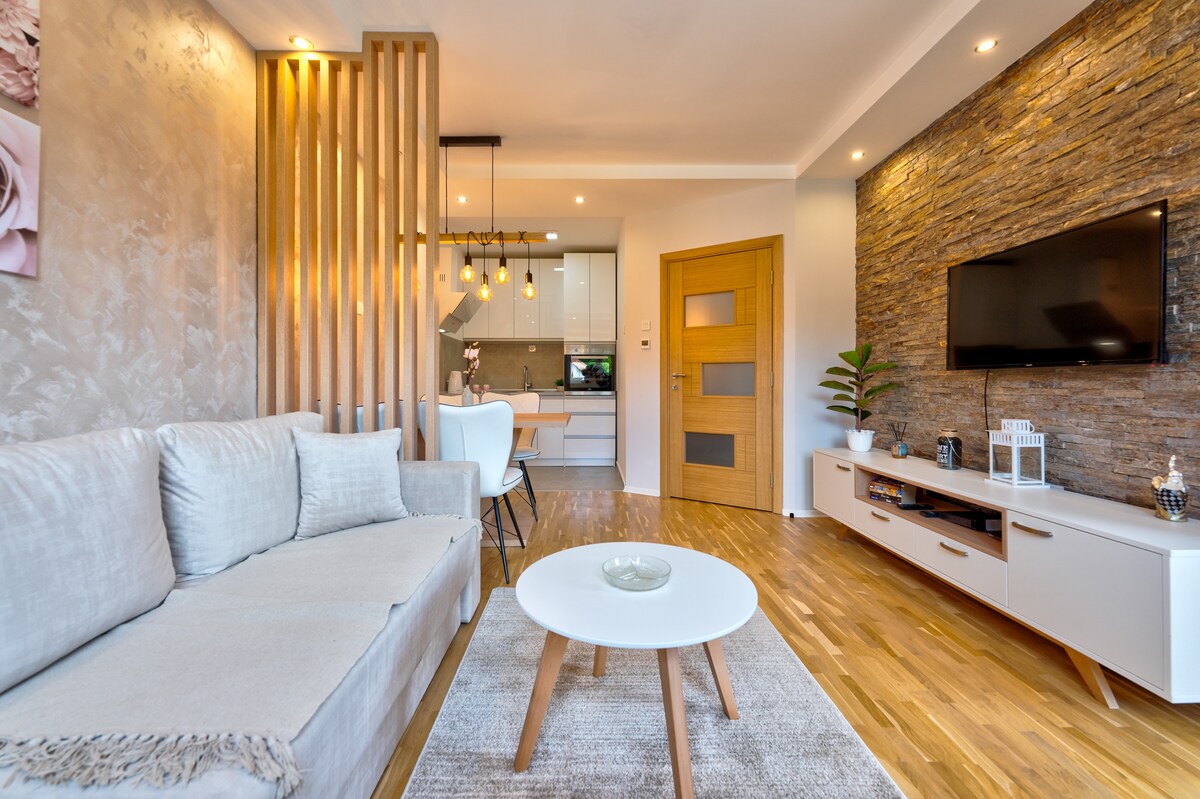 The living area features a light-colored sofa next to a round coffee table on a soft area rug. A flat-screen TV is mounted on a stone accent wall, with modern light fixtures hanging above. The open layout connects to a dining space in the background.