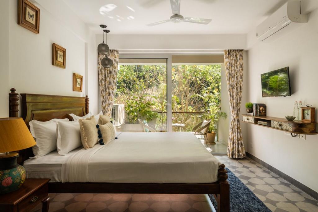 A spacious bedroom features a large bed adorned with decorative pillows. Surrounding windows invite ample natural light, while greenery is visible outside. A flat-screen TV is mounted on the wall, and a small shelf holds decorative items, creating a serene atmosphere.