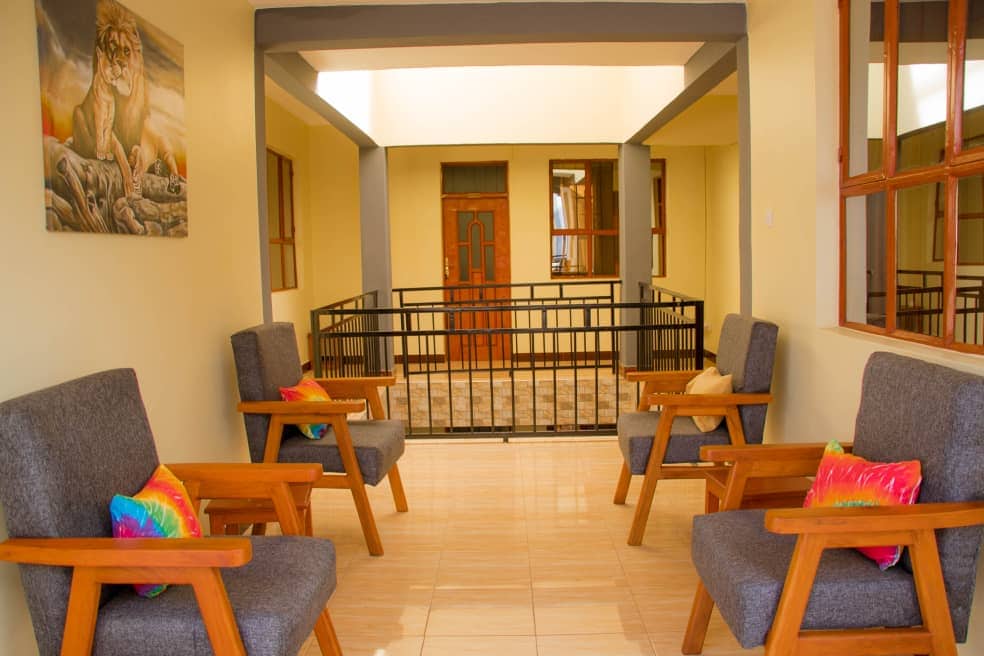 A bright corridor is lined with two comfortable seating areas, each featuring two wooden chairs with gray upholstery. Colorful throw pillows add a touch of vibrancy. The walls are painted a warm beige, complemented by natural light from surrounding windows.