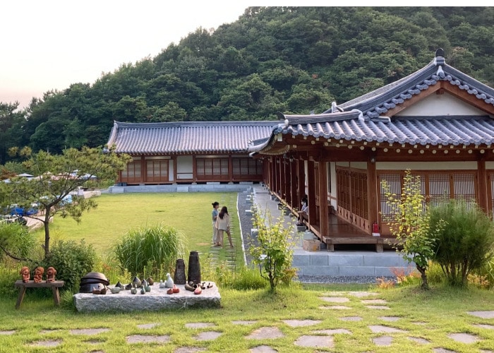 A traditional hanok is set against a lush hillside, showcasing a well-maintained garden. Stone paths lead to the wooden structure, which features a series of sliding doors. Various sculptures and planters are placed along the grassy area, emphasizing the natural ambiance.