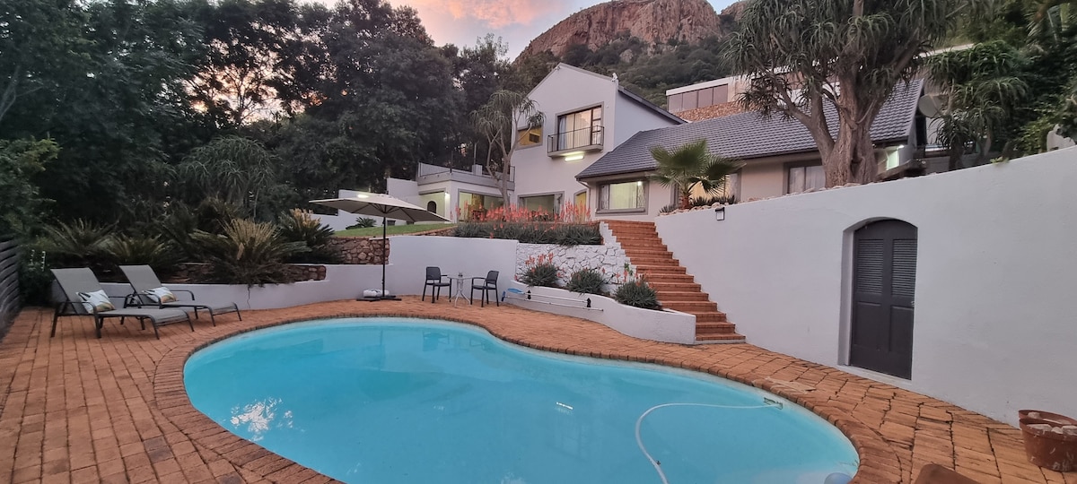 The outdoor area features a sparkling pool surrounded by a paved patio and chairs for relaxation. Lush greenery and flowering plants enhance the scenery, while the villa is visible in the background. A staircase leads towards the entrance, framed by natural landscaping.