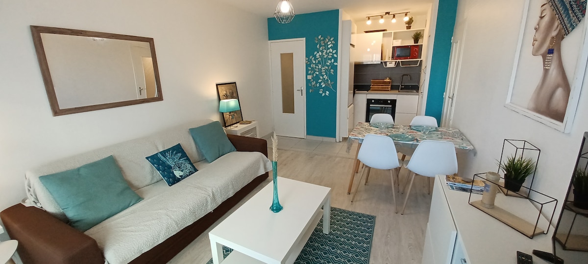 A cozy living area is featured with a light-colored sofa adorned with decorative pillows. A glass coffee table sits on a textured rug, accompanied by a dining table with white chairs. A turquoise accent wall enhances the modern decor, while a mirror reflects the ambient light.