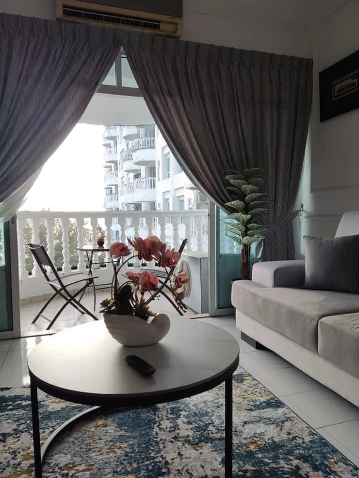 A comfortable seating area features a light gray sofa beside a round black coffee table. A vase with pink flowers sits on the table, while a colorful rug lies beneath. Large curtains frame the balcony doors, which lead to an outdoor seating arrangement.