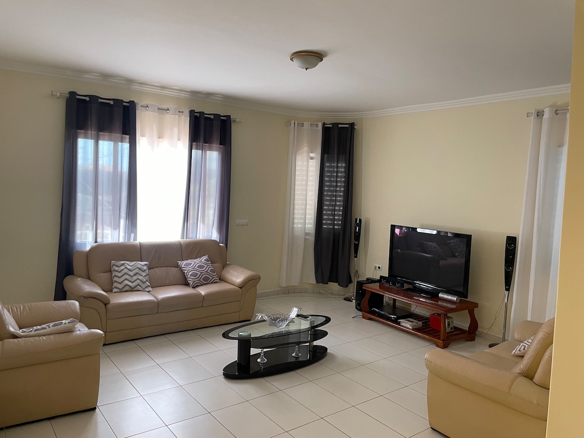 A spacious living area is furnished with two light-colored sofas and a matching armchair, arranged around a glass coffee table. A flat-screen TV is positioned on a wooden stand. Natural light enters through large windows with sheer and dark curtains.
