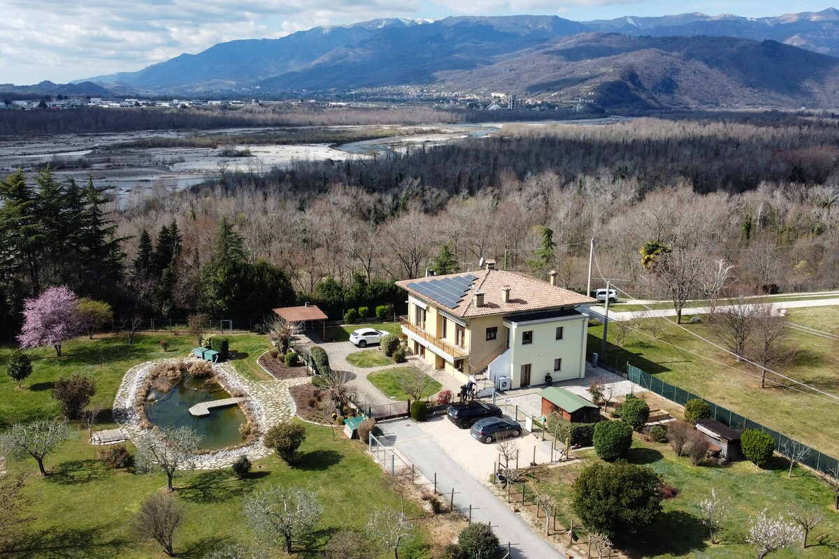 An aerial view showcases a spacious property featuring a two-story building, surrounded by gardens and greenery. A small pond is visible, with a stone border, along with several trees and parked cars. The backdrop features rolling hills and distant mountains.