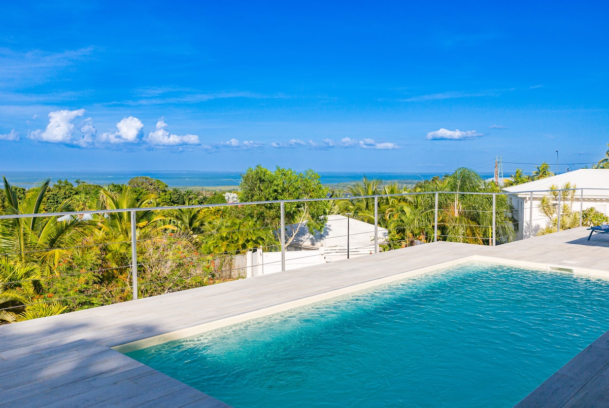 A private salt-water pool is featured, surrounded by a wooden deck with a clear view of the ocean and lush greenery. Bright blue skies enhance the panoramic scenery, creating a serene outdoor space for relaxation.
