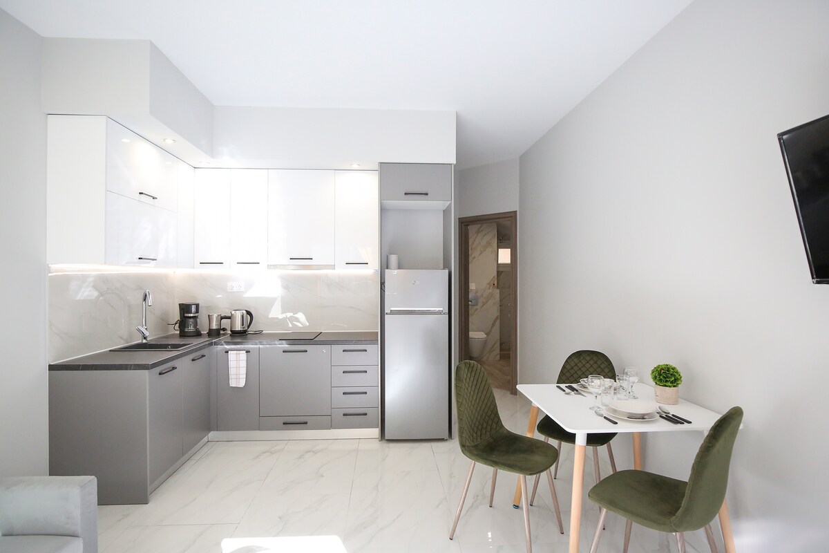 A modern kitchen area is equipped with sleek gray and white cabinets, featuring a refrigerator and a coffee maker. A small dining table is set with dishes and cutlery, accompanied by two green chairs. Light floods in from a large window, enhancing the clean design.