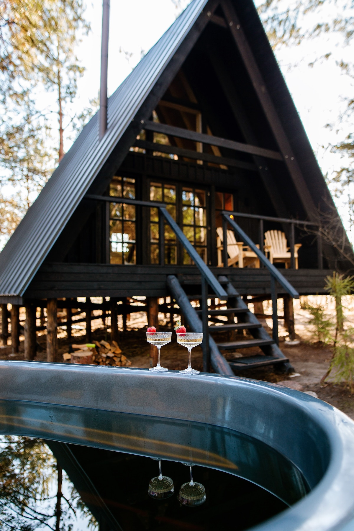 An A-frame treehouse is framed by tall pine trees, showcasing its black exterior and large windows. In the foreground, two glasses adorned with berries are placed on the edge of a hot tub, reflecting the treehouse and surroundings.