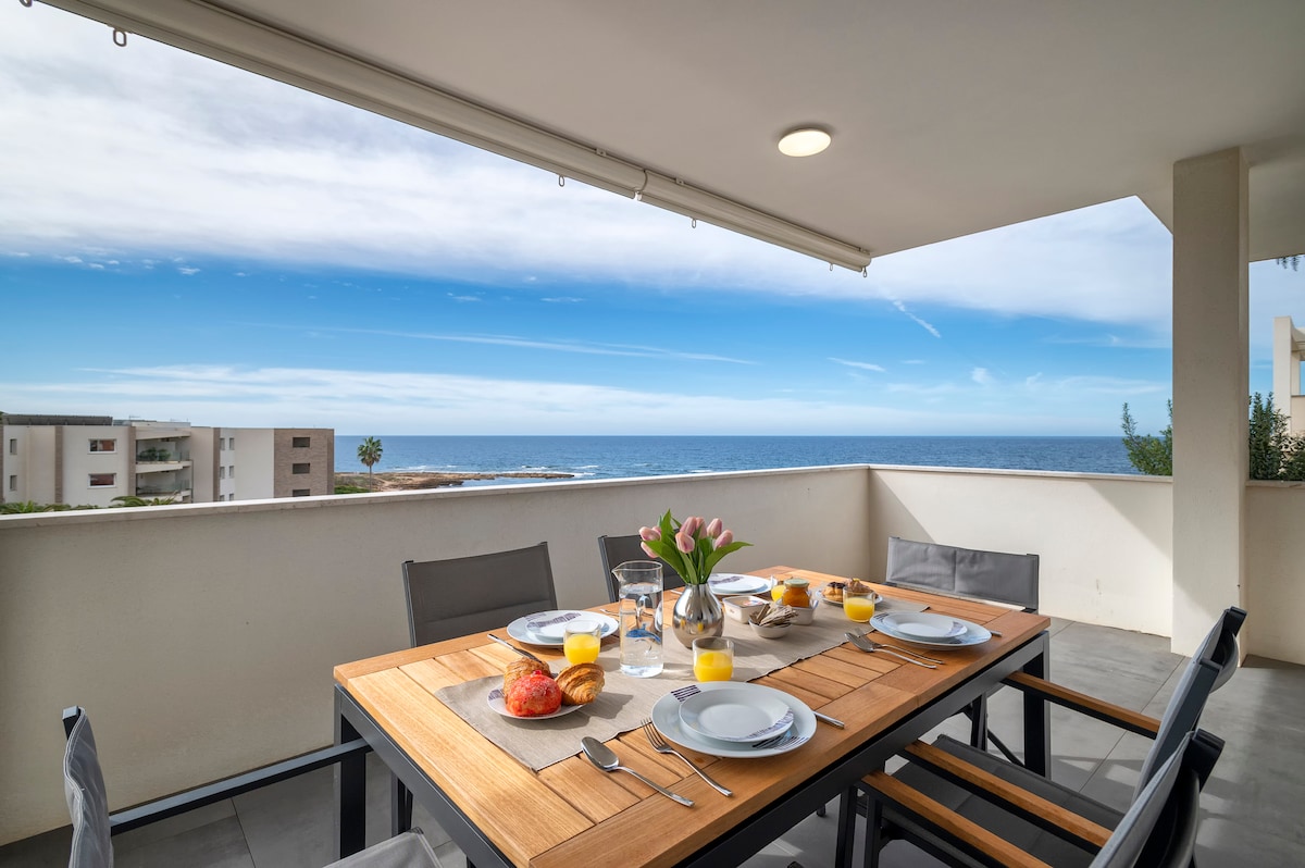 An outdoor dining space features a wooden table set for breakfast, complete with dishes, glasses, and a vase of flowers. The ocean is visible in the background, framed by a clear blue sky. Comfortable gray chairs surround the table, inviting leisurely meals.