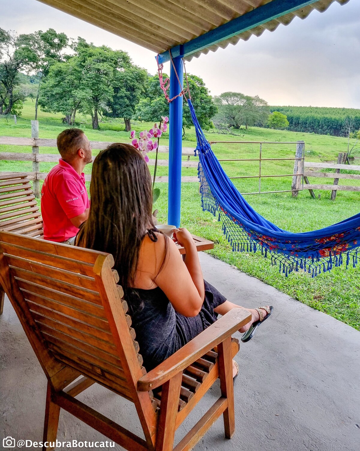 Two guests are seated on wooden chairs on a covered porch, overlooking a green landscape with trees and a wooden fence. A colorful hammock is draped nearby, creating a relaxing outdoor space. The sky is partially cloudy, adding to the serene atmosphere.