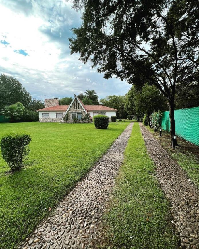 A stone pathway lined with small shrubs leads through a lush green yard, leading up to a charming house with a sloping roof and a large front lawn. Tall trees provide shade, while a vibrant green fence surrounds the property.