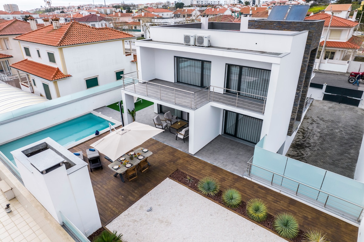 An aerial view of Villa Nazaré showcases its modern architecture, highlighting a spacious outdoor terrace with seating and a dining area. The private pool is visible beside the terrace, surrounded by wooden decking and landscaped areas, contributing to the outdoor living space.