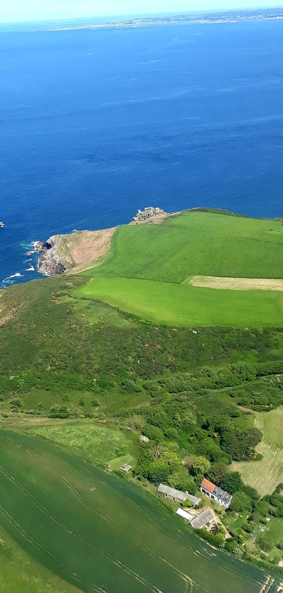 Aerial view showing a coastline with vibrant green fields meeting the blue sea. Rocky cliffs are visible along the shoreline, and the lush landscape includes patches of cultivated land, blending seamlessly into natural vegetation.