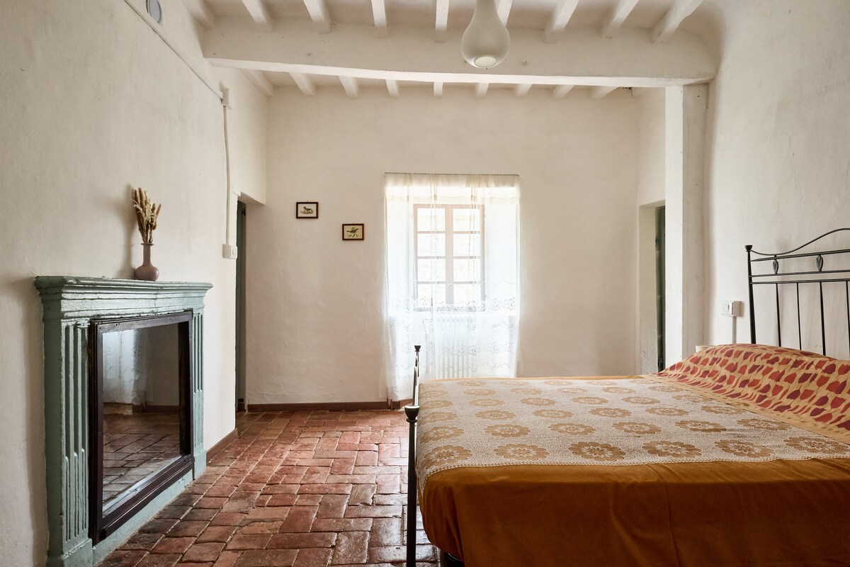 A spacious bedroom features a comfortable bed with an orange patterned blanket. A fireplace with a green mantel is positioned against a white wall. Natural light filters through a window adorned with sheer curtains, enhancing the room's airy feel.