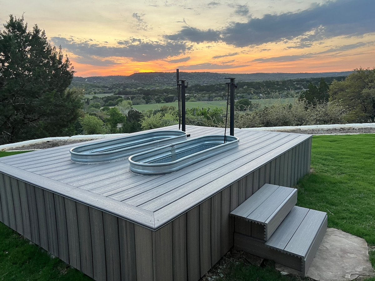 Two outdoor soaking tubs are positioned on a deck, overlooking a scenic landscape. The setting sun casts warm hues across the sky and the valleys below, creating a serene backdrop. Steps lead up to the deck, which is surrounded by grass and natural elements.