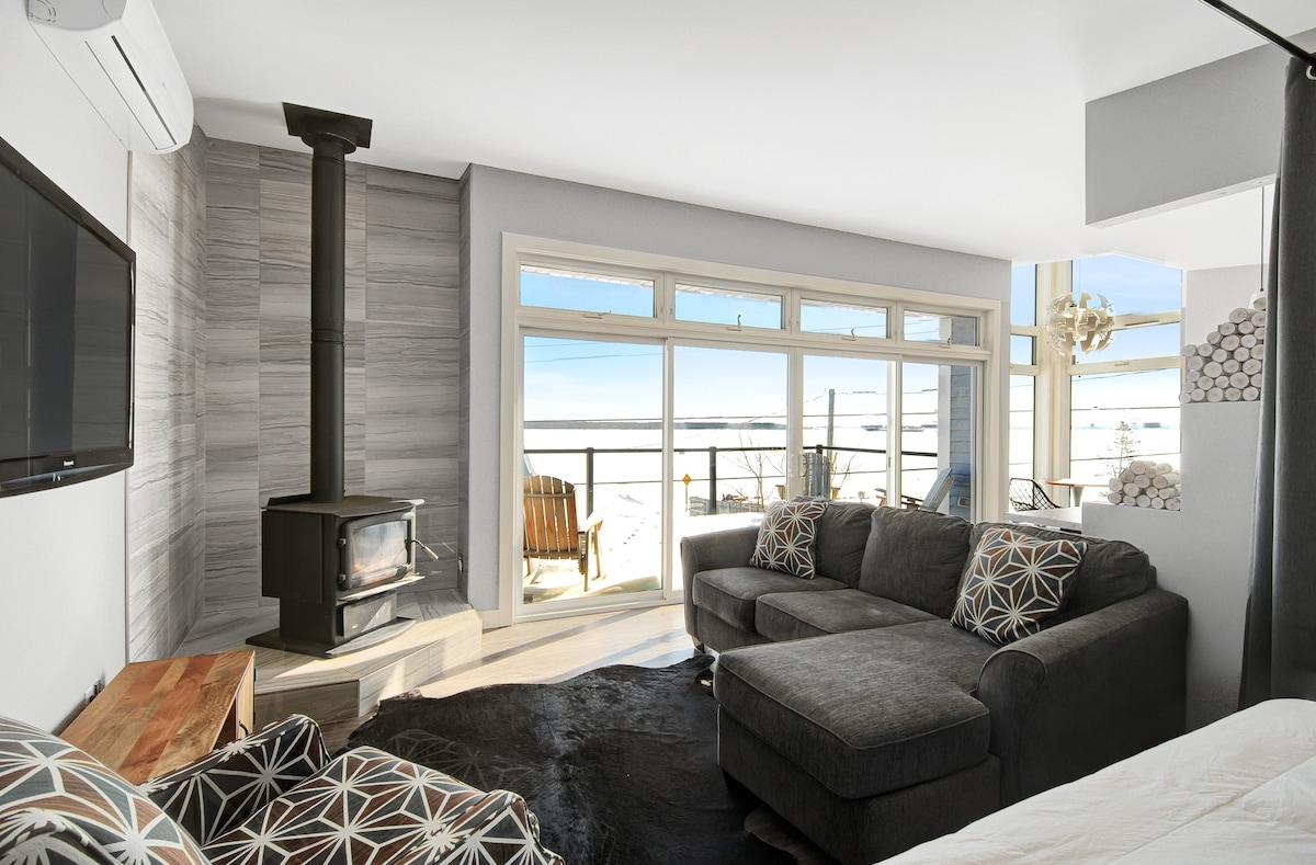 A spacious living area features a cozy seating arrangement with patterned cushions and a sleek, modern fireplace. Large windows reveal an expansive view of the ocean, allowing natural light to fill the space, while a wooden deck overlooks the tranquil surroundings.