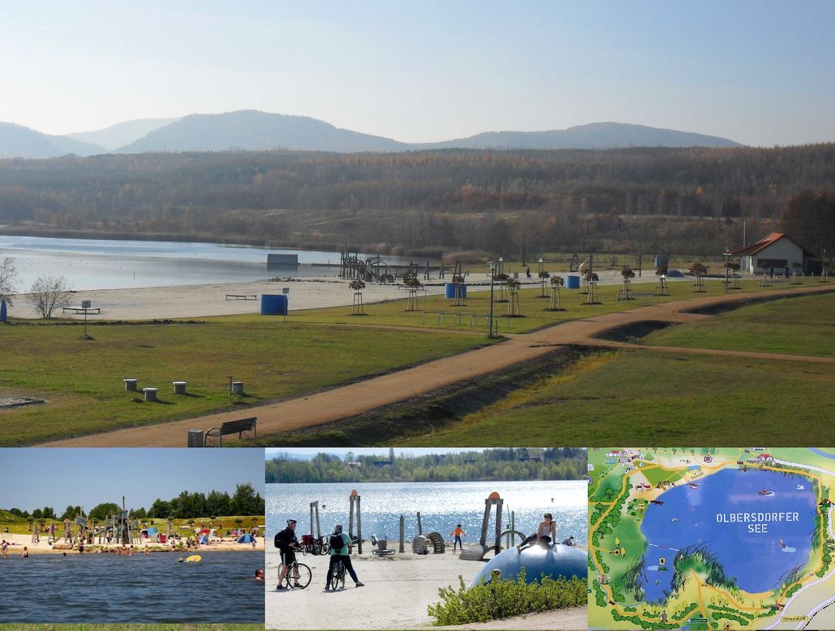A spacious lakeside area is pictured, featuring a sandy beach with distant mountains. Several benches are placed along a pathway, with recreational equipment visible. The lower section displays images of families enjoying water activities and a well-marked map of the Olbersdorfer See area.