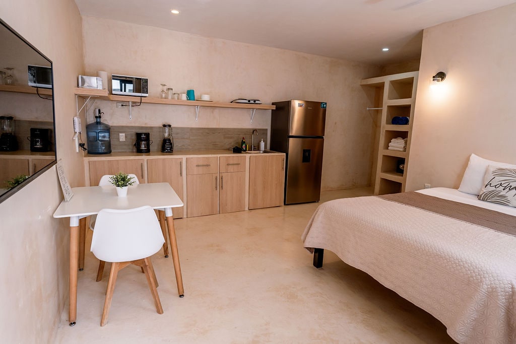 A studio space features a queen-sized bed, a small dining table with white chairs, and a kitchenette equipped with modern appliances. A sizeable wall-mounted television is positioned opposite the bed. Soft lighting and neutral-colored walls create a calming environment.